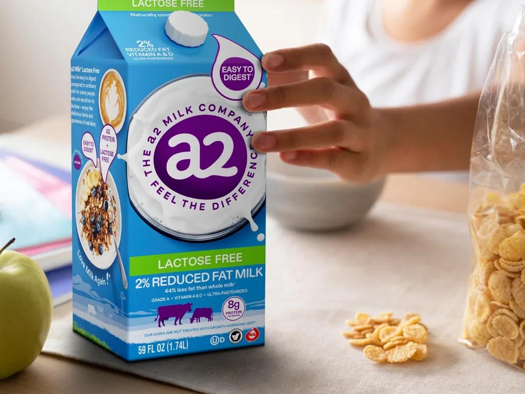 A hand reaching for a carton of a2 Milk® Lactose Free 2% Reduced Fat Milk on a breakfast table with cereal and an apple